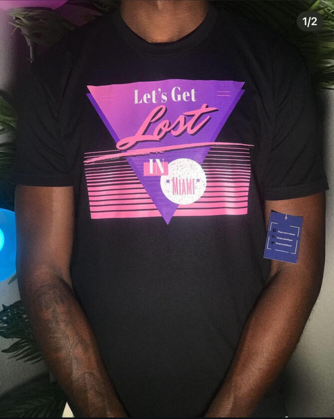 Black t-shirt featuring a purple triangle with the words “Let’s Get Lost in Miami” in retro 80s-style font, pink horizontal stripes, and pastel highlights representing a vintage South Beach aesthetic.