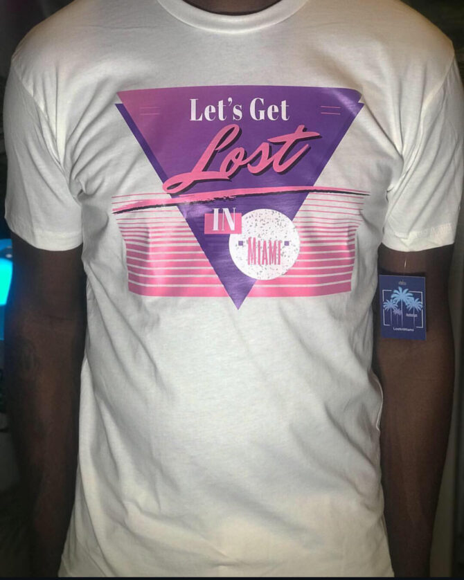 White t-shirt with a purple triangle, pink neon lines, and the phrase “Let’s Get Lost in Miami” in a bold retro-futuristic font, reflecting 80s-inspired Miami fashion.